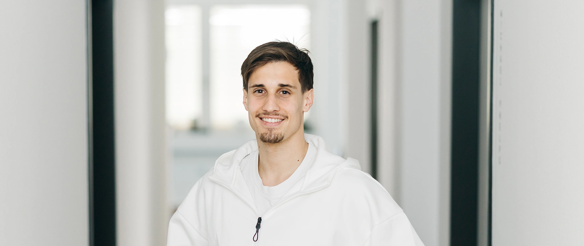 Porträt eines lächelnden, jungen männlichen Physiotherapeuten im weißen Hoodie in den hellen Fluren der Praxis David Lucksch.