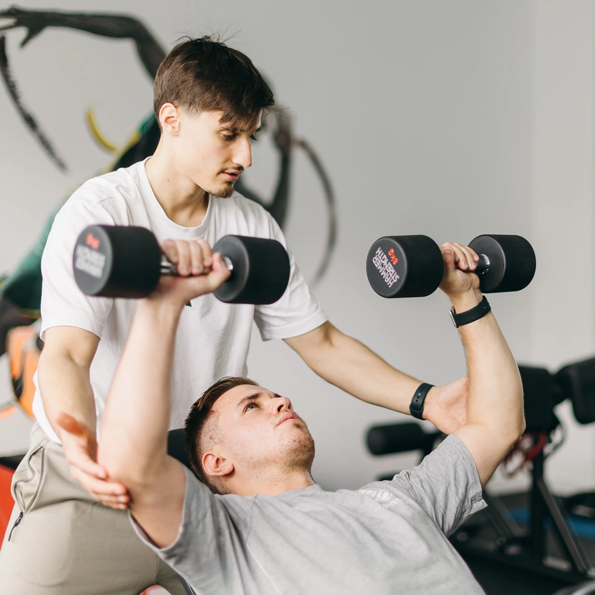 philosophie05 Sportphysiotherapeut korrigiert die Ausführung beim Kurzhantel-Bankdrücken während einer Trainingseinheit bei DuoLevitas.